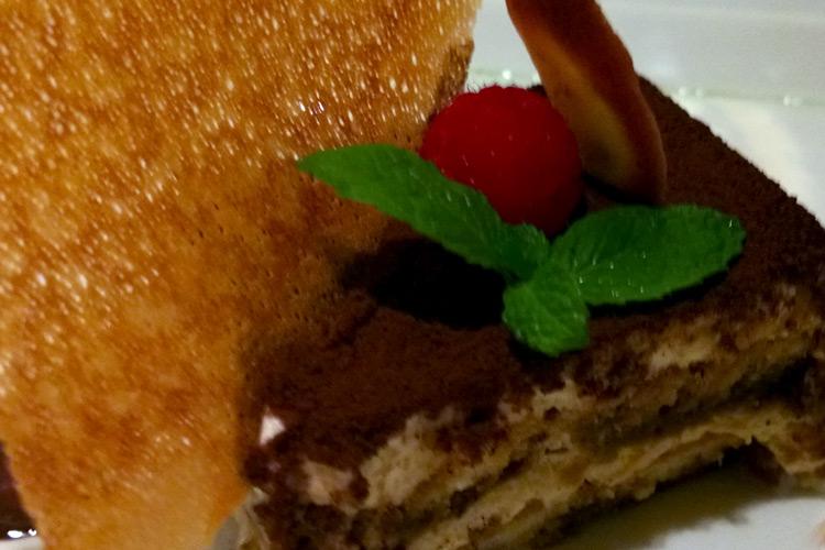 U trozo de tiramisu decorado con cacao, menta y frambuesa, acompañado con barquillo.