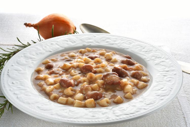Un plato blanco de loza con pasta y alubias, una cebolla y una cuchara al fondo.