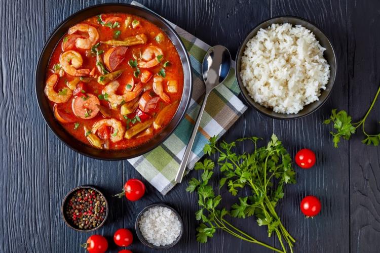 Gumbo de gambas y salchicha con un cuenco de arroz blanco y condimentos sobre pizarra.