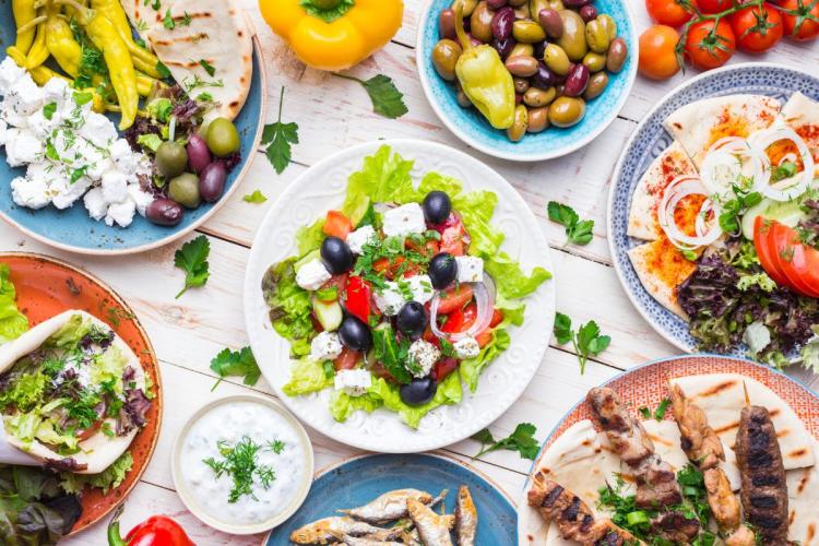 Una selección de platos de la cocina griega, incluyendo meze, gyros, souvlaki, pan pita, ensalada griega, tzatziki, queso feta, aceitunas.