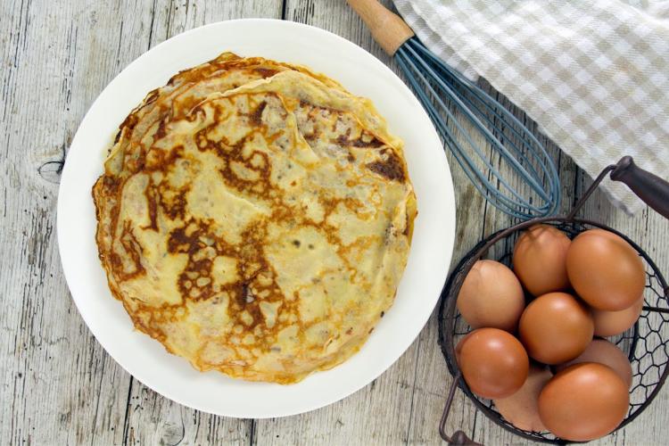 Un palto con crêpes caseras con un batidor de globo y un cuenco con huevos frescos al lado.