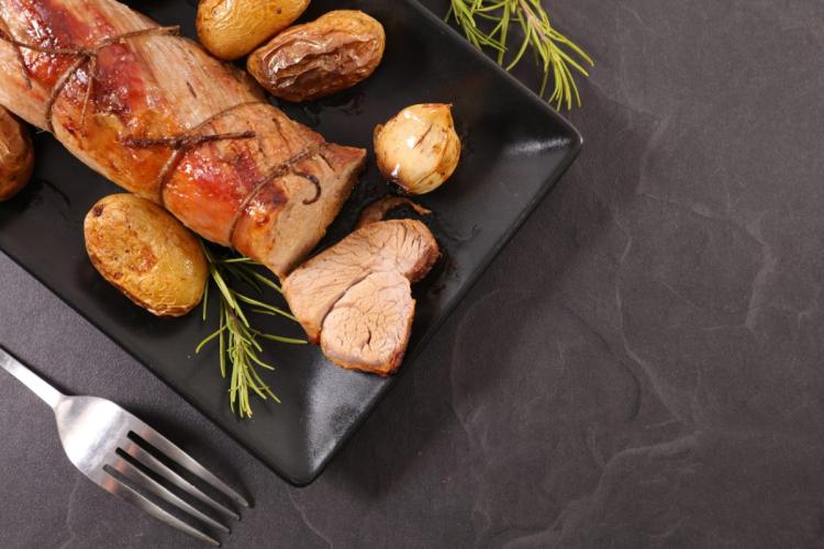 Una fuente de color negro con un solomillo de cerdo al horno con patatas asadas y hierbas.