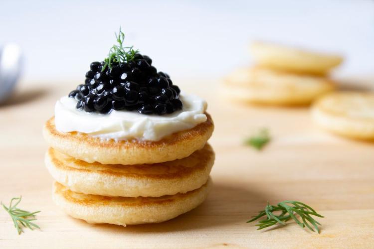Tres blinis apilados, cubiertos con nata agria y caviar negro, adornados con una ramita de eneldo.