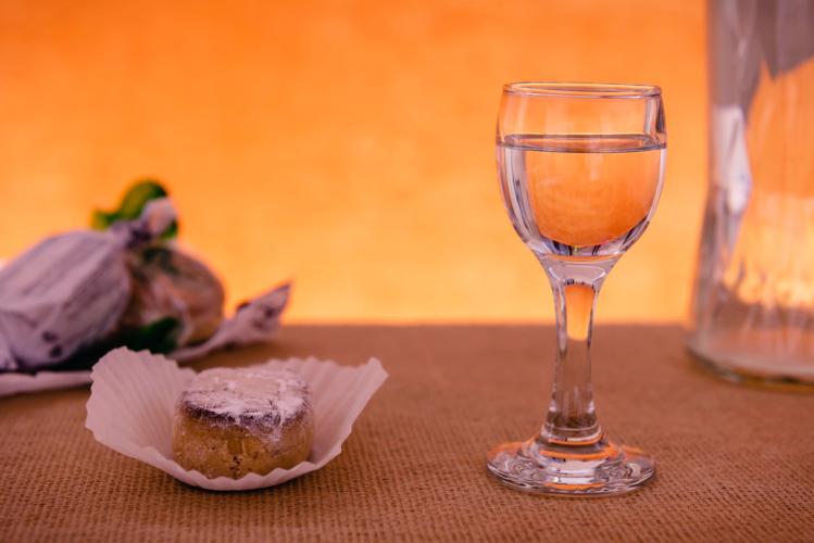 Una copa de licor de anís transparente junto a un polvorón, un dulce tradicional español, en un envoltorio de papel blanco sobre una superficie de arpillera, con un fondo naranja cálido.