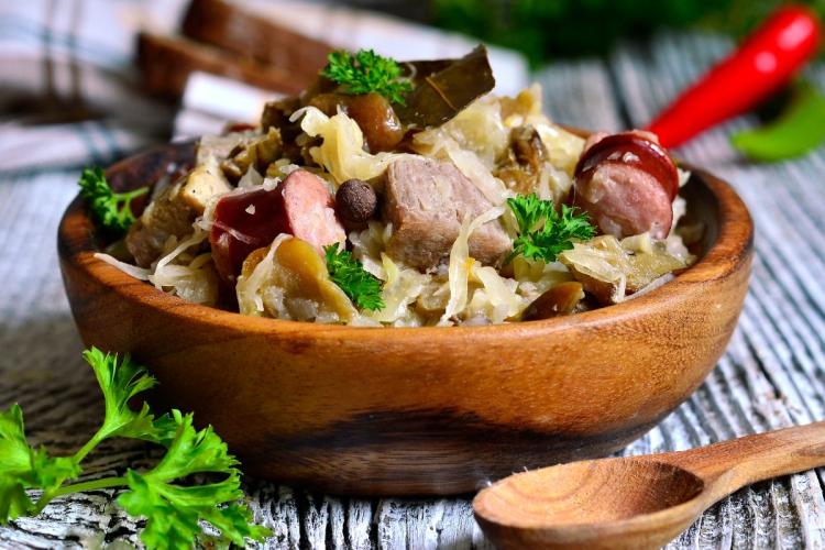 Bigos tradicional polaco servido en un cuenco rústico de madera sobre una mesa de madera desgastada, con chucrut, trozos de cerdo, rodajas de salchicha, champiñones y adornado con perejil fresco.