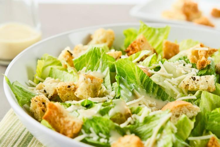 Una ensalada César en su ensaladera con un poco de aliño César en un recipiente al fondo.