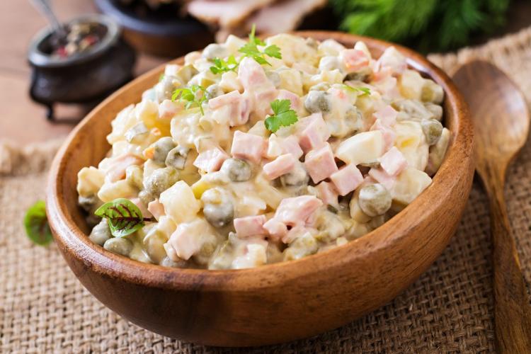 Un cuenco de madera con ensalada Olivier, o ensalada rusa, con patata, zanahoria, guisantes, jamón y pepinillos troceado y mezclados con mayonesa. El cuenco está colocado sobre una superficie de arpillera y hay una cuchara al lado.