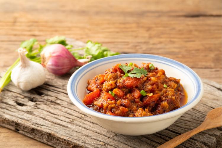 Un cuenco de nam prik ong, una salsa de chile tailandesa, sobre una superficie de madera rústica con ajo, chalotes y cilantro en el fondo.