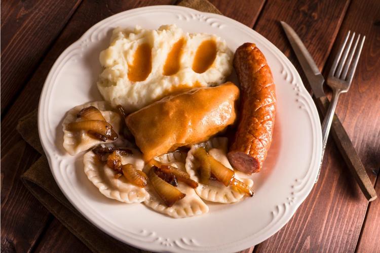 Plato con comida tradicional polaca que incluye pierogies con cebolla salteada, puré de papas con salsa, salchicha kielbasa y un rollo de repollo relleno cubierto con salsa.