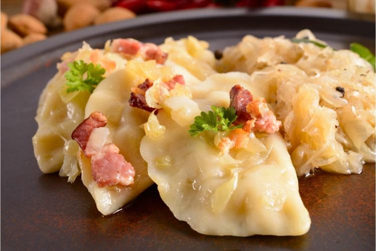 Plato con tres pierogies cubiertos con cebolla salteada y tocino, servido con chucrut.