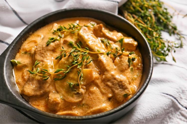 Un plato de carne Stroganoff adornada con tomillo fresco, colocado sobre un paño blanco con más hierbas aromáticas al fondo