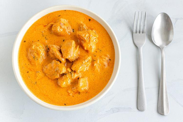 Un recipiente con pollo a la mantequilla en su salsa. Hay un tenedor y una cuchara al lado derecho.