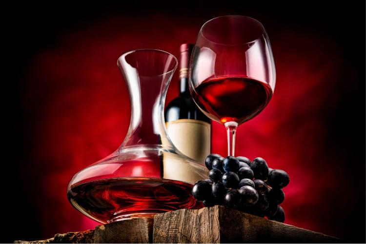 Un decanador con vino tinto, una copa de vino tinto y un racimo de uvas con una botella de vino al fondo.