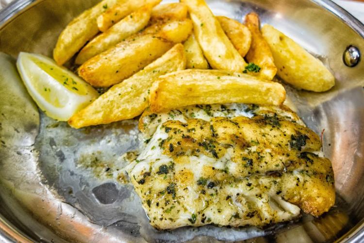 Filete de kingklip, un pescado de Namibia, a la parrilla condimentado con hierbas, servido junto con papas fritas doradas cortadas gruesas y una rodaja de limón en un plato de metal.