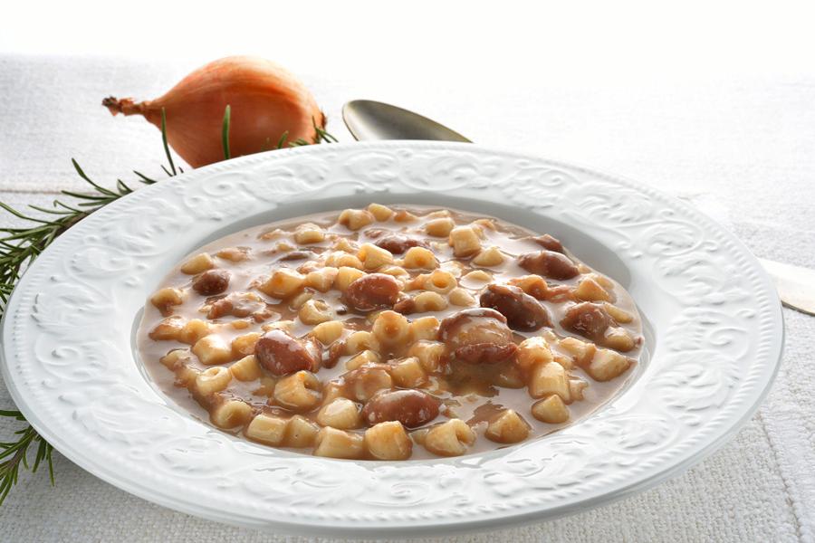 Un plato blanco de loza con pasta y alubias, una cebolla y una cuchara al fondo.