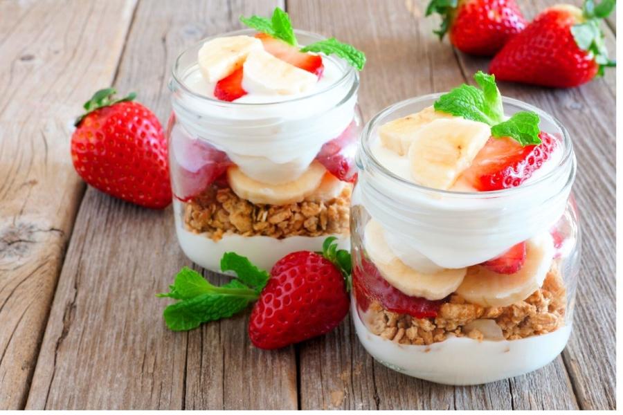 Copas de fruta, yogur y granola, una con plátano y otra con fresa, en tarros de vidrio.