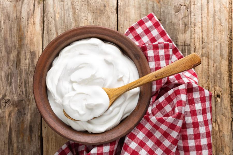 Un cuenco de madera con yogur natural con una cuchara de palo sobre una servilleta de cuadros en una mesa de madera.