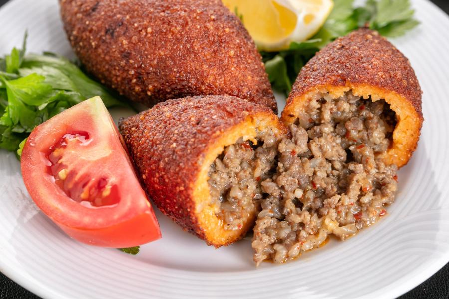 Plato con croqueta de kibbeh partida por la mitad, mostrando su relleno de carne picada sazonada, acompañado de una rodaja de tomate, perejil y un cuarto de limón.