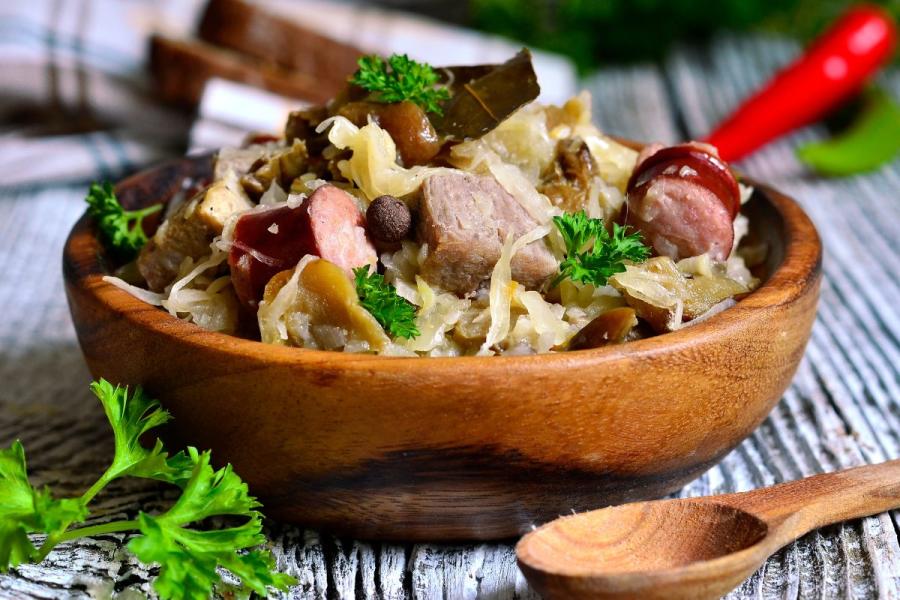 Bigos tradicional polaco servido en un cuenco rústico de madera sobre una mesa de madera desgastada, con chucrut, trozos de cerdo, rodajas de salchicha, champiñones y adornado con perejil fresco.
