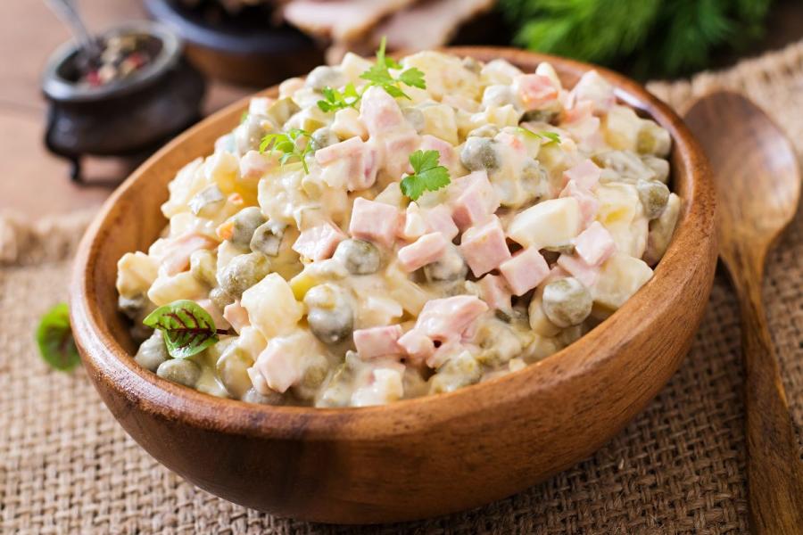 Un cuenco de madera con ensalada Olivier, o ensalada rusa, con patata, zanahoria, guisantes, jamón y pepinillos troceado y mezclados con mayonesa. El cuenco está colocado sobre una superficie de arpillera y hay una cuchara al lado.