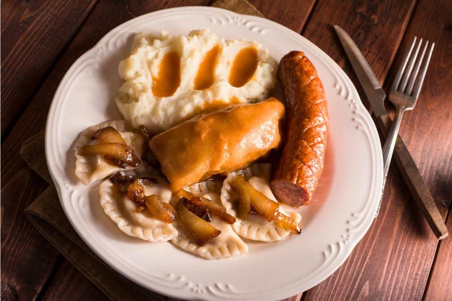 Plato con comida tradicional polaca que incluye pierogies con cebolla salteada, puré de papas con salsa, salchicha kielbasa y un rollo de repollo relleno cubierto con salsa.