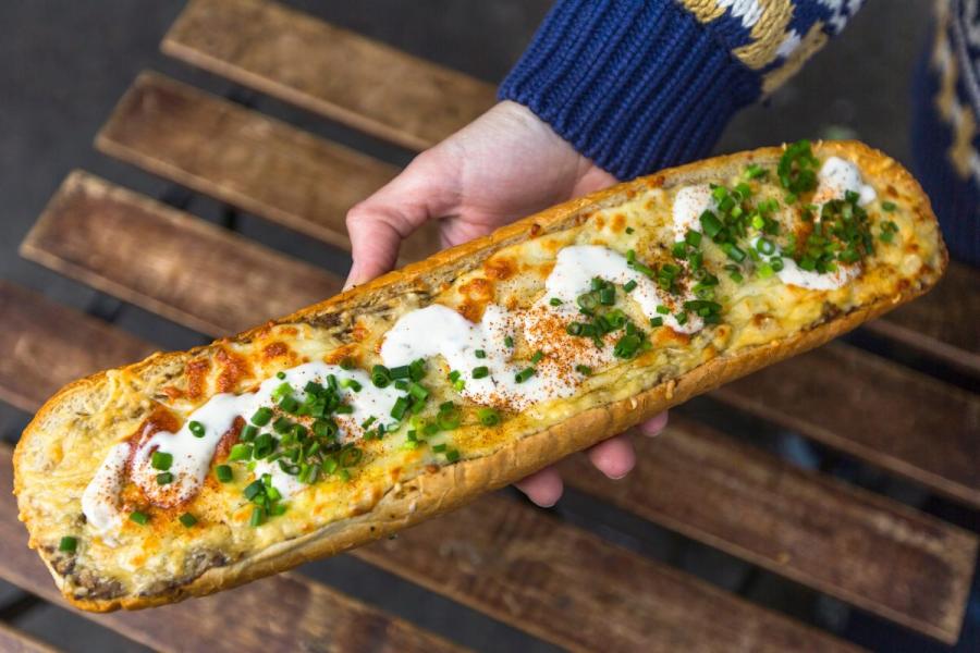 Imagen de una zapiekanka, una comida callejera polaca, con queso derretido, champiñones salteados, crema agria y cebolla verde picada. Una mano sostiene el sándwich.
