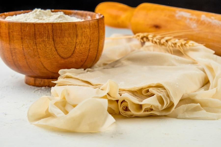 Masa filo casera con un cuenco de harina y un rodillo de madera detrás.