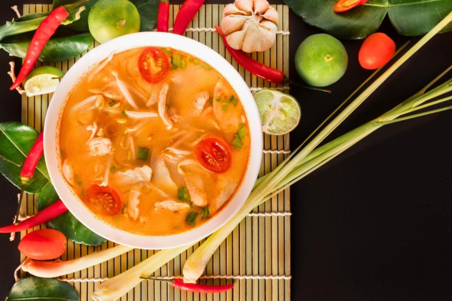 Un bol des sopa tom yum gai con pollo, tomates cereza y hierbas, rodeado de ingredientes frescos como hierba de limón y limas.