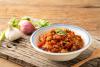 Un cuenco de nam prik ong, una salsa de chile tailandesa, sobre una superficie de madera rústica con ajo, chalotes y cilantro en el fondo.